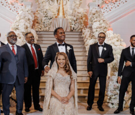 A woman in a gold gown sits as a man stands behind her on a floral staircase with men in suits and gold monogram initials above.