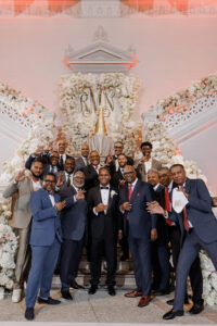 Groom poses with a large group of guests in tuxedos and suits in front of the lavish floral staircase and wedding cake display.