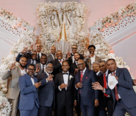 Men in suits smile and celebrate on flower-lined steps with a gold monogram at a formal Louisiana wedding.