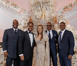 Five people in formal attire smile before white florals and a gold monogram at an elegant New Orleans wedding with grand staircases.