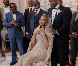 Bride in ornate gown and groom in tuxedo smile, surrounded by guests at a beautifully decorated indoor wedding venue.