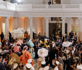 Indoor Louisiana-style wedding with a brass band, feathered dancers, and guests celebrating in an elegant columned hall.