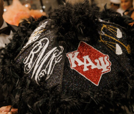 Person in a tuxedo holds a black feathered umbrella with white initials, gold designs, and red ΚΑΨ patch—stylish New Orleans wedding inspo.