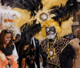 Man in black and yellow feathered headdress, animal print, and face paint at indoor Louisiana wedding celebration.