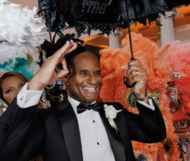 Man in tuxedo smiles, holding a black feathered umbrella amid colorful costumes at a vibrant New Orleans wedding.