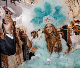 Woman in a light blue feathered costume and headdress smiles at an indoor event as guests take photos and hold feathered fans.