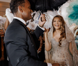 Man in tuxedo with black feathered umbrella and smiling woman in ornate gown at festive New Orleans wedding indoors.