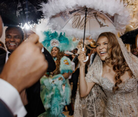 Bride in ornate gown smiling with decorated umbrella, surrounded by dancing guests at vibrant New Orleans wedding.