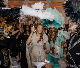 A joyful bride in a sparkling dress is surrounded by guests in elaborate costumes at a lively New Orleans wedding.