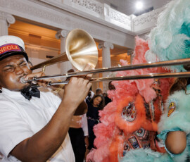 Musician in white uniform and Kinfolk hat plays trombone beside vibrant feathered guests at ornate New Orleans-inspired wedding.