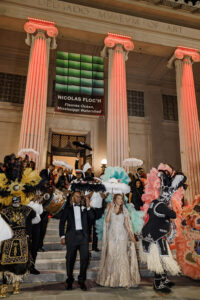 Bride and groom lead a vibrant second line celebration outside the museum surrounded by Mardi Gras Indians, parasols, and brass band musicians.