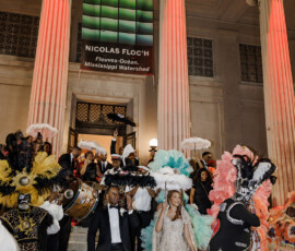 A couple leaves a museum amid musicians and Mardi Gras costumes under red-lit columns, inspiring Gulf South weddings.