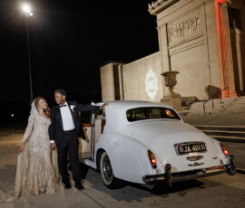 A couple in formal wear stands by a white vintage car at night before a grand, lit building—Gulf South wedding inspiration.