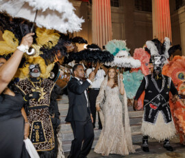 People in feathered costumes and formal wear parade with parasols up stone steps at night before tall pillars, celebrating New Orleans-style.