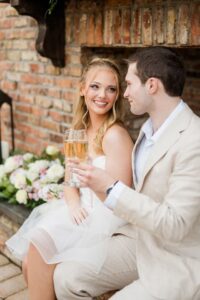 Smiling couple in light attire with champagne at their Gulf South wedding, by a brick wall and floral arrangement.