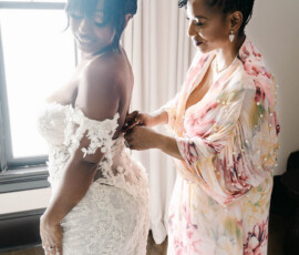 Bride in white lace gown smiles as woman in floral robe zips her dress by sunny window before Louisiana wedding ceremony.