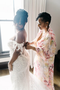 Bride smiles while a woman in a floral robe buttons the back of her lace off-the-shoulder wedding gown beside a bright window.
