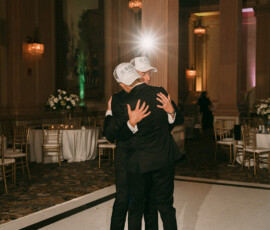 Two people in suits and white hats hug on a wedding dance floor, elegant tables and columns around them, bright light behind.