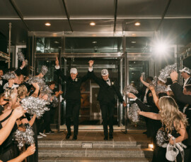 Two people in suits and white caps celebrate outside a building, holding hands as a joyful crowd waves silver pom-poms.