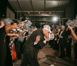 Two grooms in suits and white caps kiss as guests cheer with silver pom-poms outside a building at their Gulf South wedding.