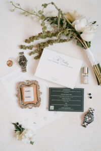 Flat lay of wedding invitations, watches, rings, perfume, pendant, bouquet, and accessories on a light surface.