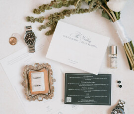 Flat lay of wedding invitations, watches, rings, perfume, pendant, bouquet, and accessories on a light surface.