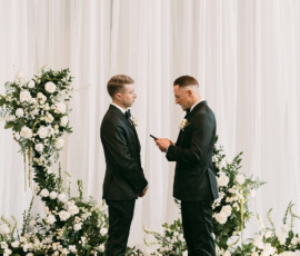Two men in black suits face each other at a Louisiana wedding, surrounded by white flowers as one reads from his phone.