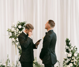 Two men in tuxedos share an emotional moment at a Gulf South wedding, with white flowers and draped curtains behind them.