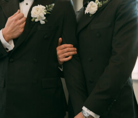 Two men in black tuxedos link arms and hold hands, sharing a tender moment at their Louisiana wedding.