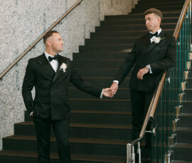 Two men in tuxedos hold hands on a Louisiana wedding staircase, gazing at each other beside a granite wall and metal railing.