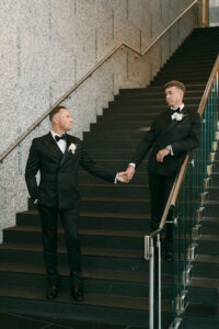Two men in tuxedos hold hands on a Louisiana wedding staircase, gazing at each other beside a granite wall and metal railing.