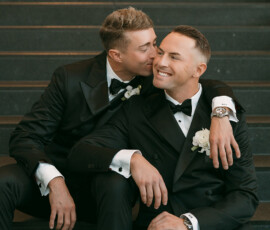 Two men in black tuxedos share a happy, affectionate moment on stairs at their elegant New Orleans wedding.