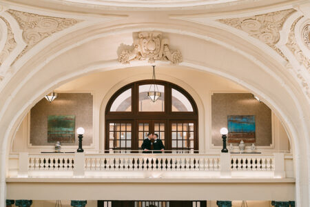 Couple stands on ornate balcony in grand building with arched ceilings, reflecting a timeless New Orleans wedding.