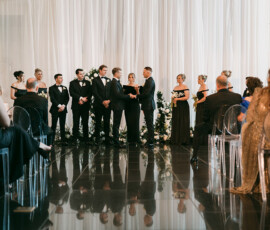 Louisiana wedding ceremony with wedding party at the altar, officiant center, guests on clear chairs, white drapes and flowers behind.