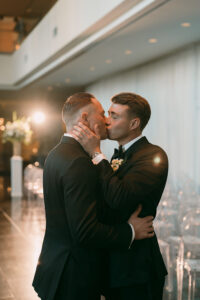 Two men in suits kiss and embrace at a beautifully lit wedding ceremony, with empty chairs in the background.