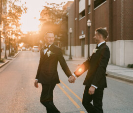 Two men in suits hold hands and smile at sunset on a city street, sunlight shining between them—a joyful wedding moment.