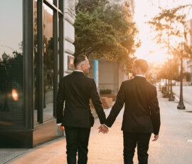 Two men in suits hold hands on a city sidewalk at sunset, with buildings behind—romantic Louisiana wedding inspiration.