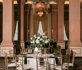 Elegant New Orleans wedding table with white cloth, gold chairs, floral centerpiece, columns, and chandeliers.