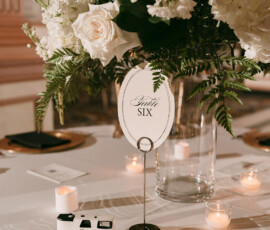Elegant white table with floral centerpiece, Table Six sign, candles, camera, and card for wedding or New Orleans ceremony.