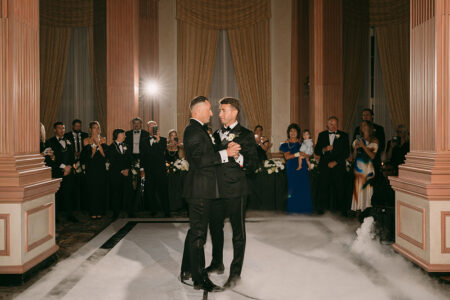 Two grooms in tuxedos share a first dance amid elegant columns, draped curtains, and soft lighting at their wedding reception.