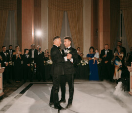 Two grooms in tuxedos share a first dance amid elegant columns, draped curtains, and soft lighting at their wedding reception.