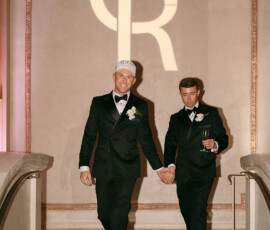 Two men in black tuxedos, one in a white cap, hold hands and smile at their New Orleans wedding with a CR monogram behind them.