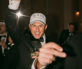 Man in tuxedo and TIL DEATH hat smiles at lively New Orleans wedding, holding drink and glowing foam stick among formal guests.