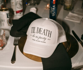White cap with “TIL DEATH do us party” on gold charger plate, black napkin, and drinks—unique Louisiana wedding reception inspo.