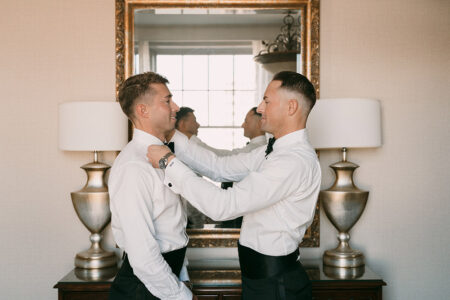 Two men in formal wear stand by a mirror as one adjusts the other’s bow tie before a Louisiana wedding.