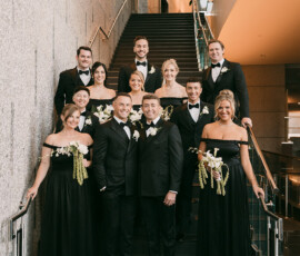 A joyful wedding party poses on an indoor staircase, with six women in black dresses and six men in tuxedos, all smiling.