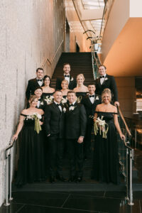 A joyful wedding party poses on an indoor staircase, with six women in black dresses and six men in tuxedos, all smiling.