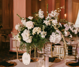 Elegant wedding table with tall white floral centerpiece, gold chairs, candles, plates, and a QR code in a softly lit venue.
