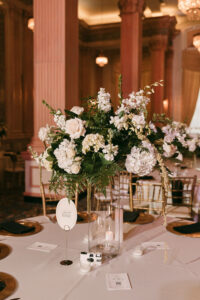 Elegant wedding table with tall white floral centerpiece, gold chairs, candles, plates, and a QR code in a softly lit venue.