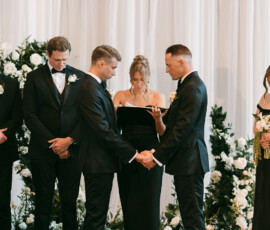 Two grooms in black suits hold hands at their Louisiana wedding, with attendants, white flowers, and draped curtains behind them.